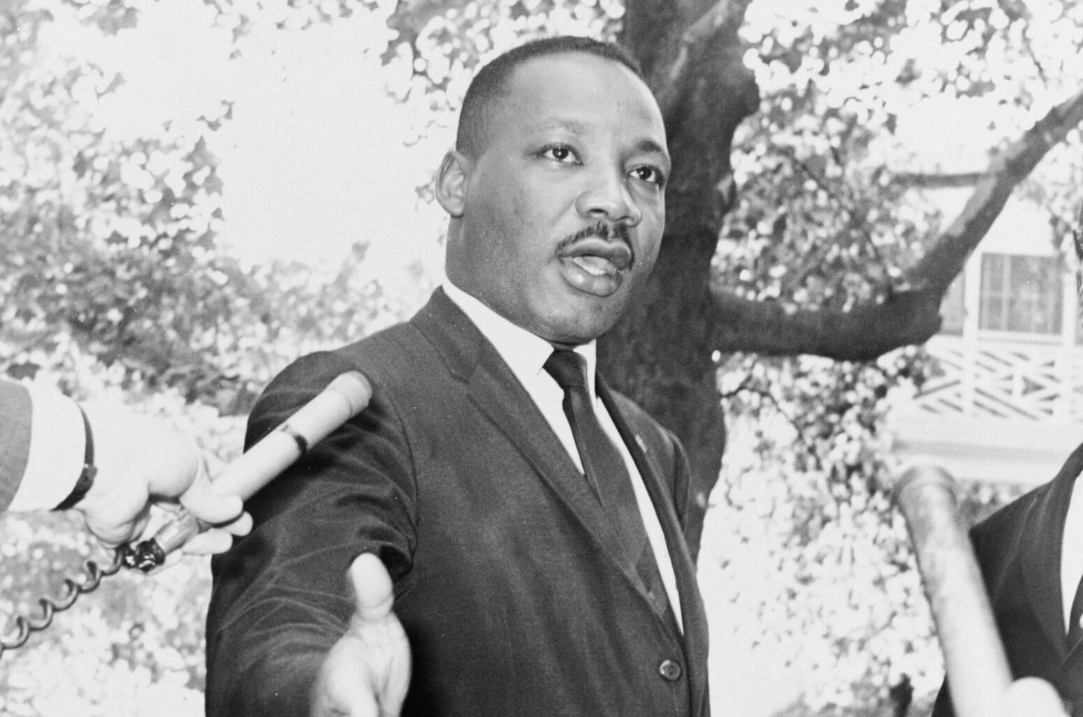A black-and-white photo of Martin Luther King Jr. He is talking to someone beyond the camera, and holding his hand out. He is standing outside, and people hold microphones up to him.