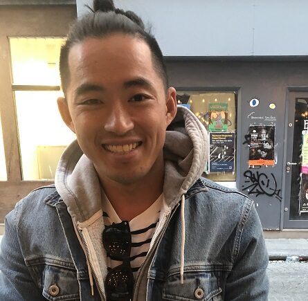Patrick J. Zhou, an Asian American man, sits at an outdoor table. He's wearing a denim jacket, gray hoodie, and striped white-and-black shirt with sunglasses tucked at the neck. There's a street and storefront behind him.