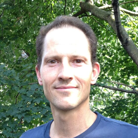 A white man with close-cropped dark hair. He's wearing a blue T-shirt and standing under a tree