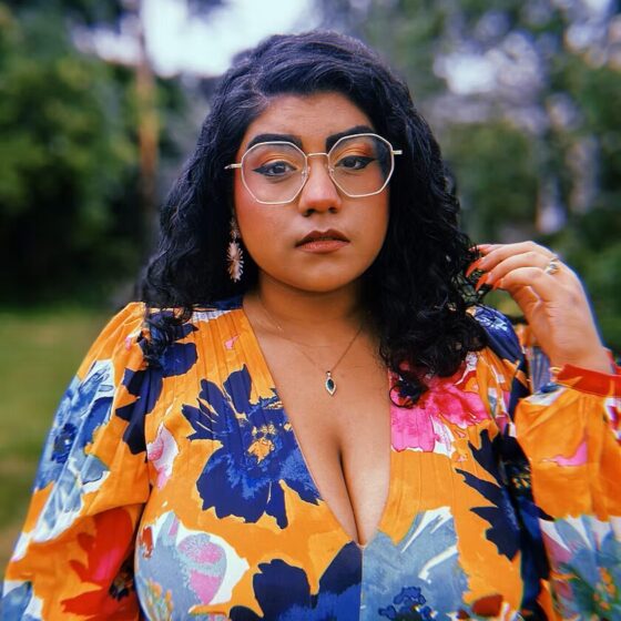 An Indo-Guyanese woman with dark curly hair and glasses. She's wearing an orange, blue, and pink blouse and is standing outside near trees.
