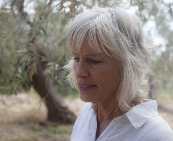 A woman with white hair, wearing a white collared shirt. She's looking down and at the left without much expression. There's a tree behind her.