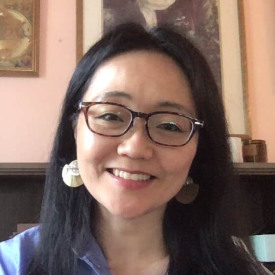 An Asian American woman with long dark hair, red glasses, and danging earrings. She's wearing a button-up blue shirt and sitting in front of a brown horizontal bookshelf and pink wall with art on it