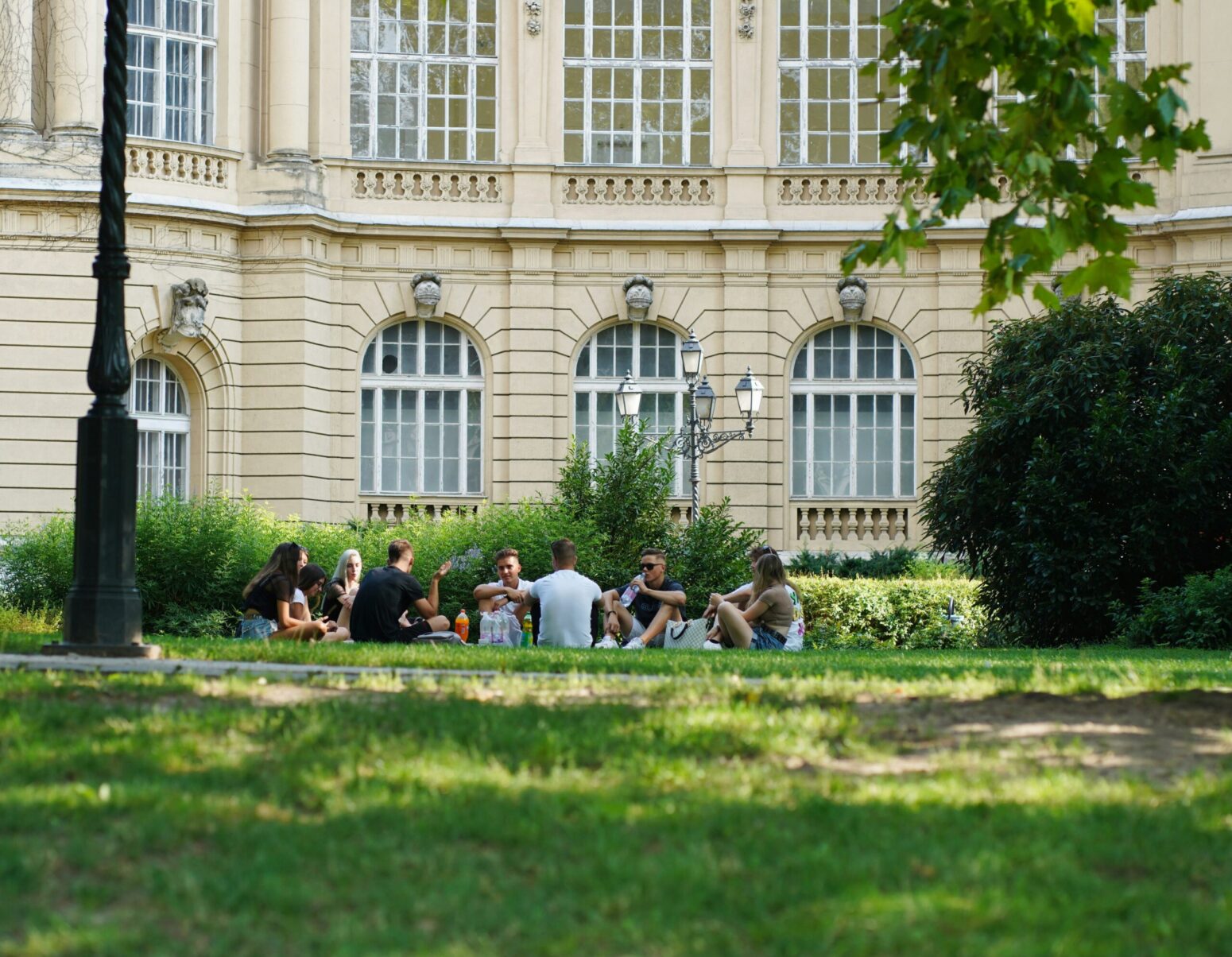 A group of young adults sitting in a circle on some grass in a mix of sunlight and shade, with a white stately building behind them
