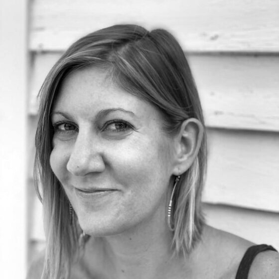 A black-and-white photo of a white woman with shoulder-length light hair. She's smiling, wearing a dark tank top, and standing near a building's siding.