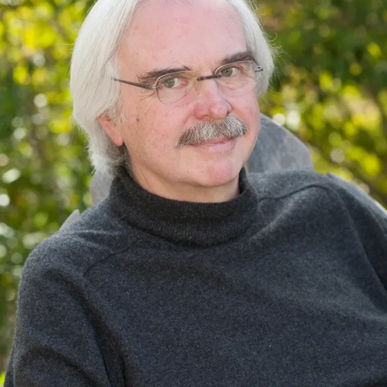 A man with white hair and a gray mustache. He's wearing glasses and a gray turtleneck and is outside in front of greenery