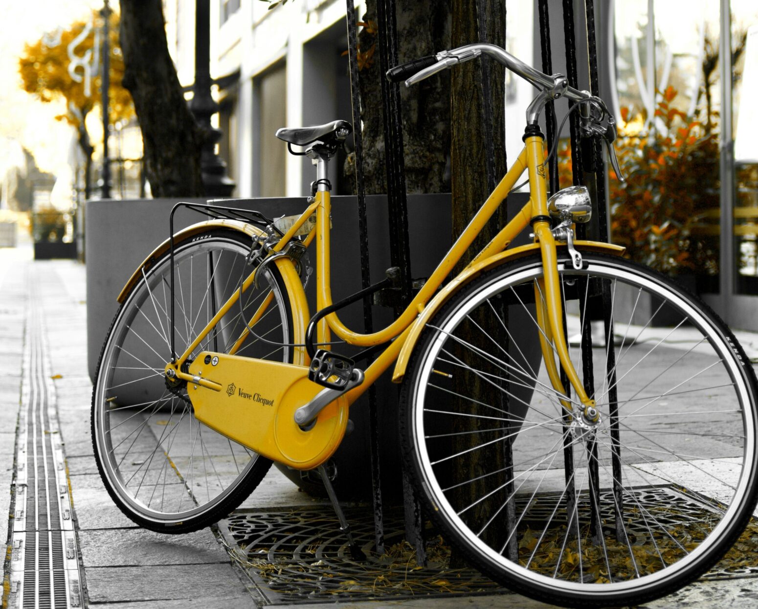 A bicycle with a yellow body leaning against a tree with a protective metal frame, in an urban setting