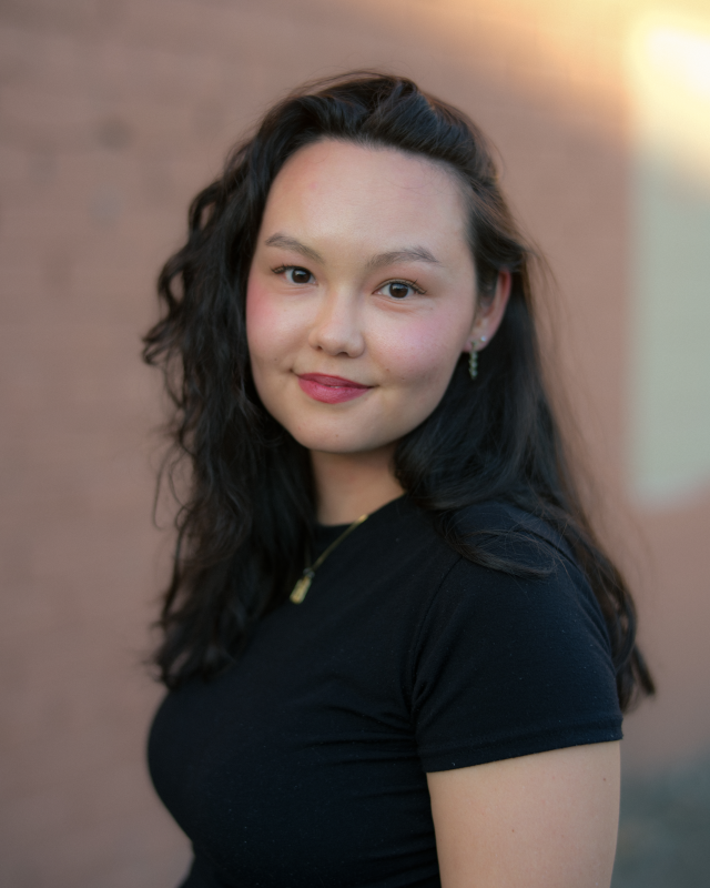Arah Ko, an Asian-American woman with dark curly hair cascading down her shoulders, smiles in front of a tan background. She wears a black tee shirt.