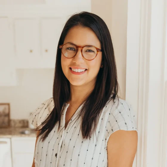 A Ecuadorian woman with brown hair past her shoulders. She's standing in a room with white cabinets and beige walls, wearing a button-down white blouse and brown glasses.