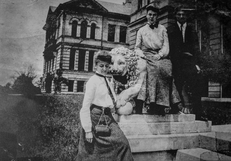 Three Edwardian youths--two women and a man--sit on a stone lion on the University of Cincinnati campus.