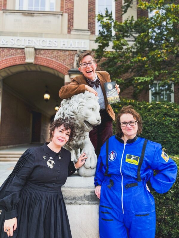 Three white women posing with a stone lion on the UC campus. One wears a bow tie, glasses, and a corduroy jacket, one wears a blue space suit and glasses, and one wears a black Victorian-style dress and Victorian jewelry.