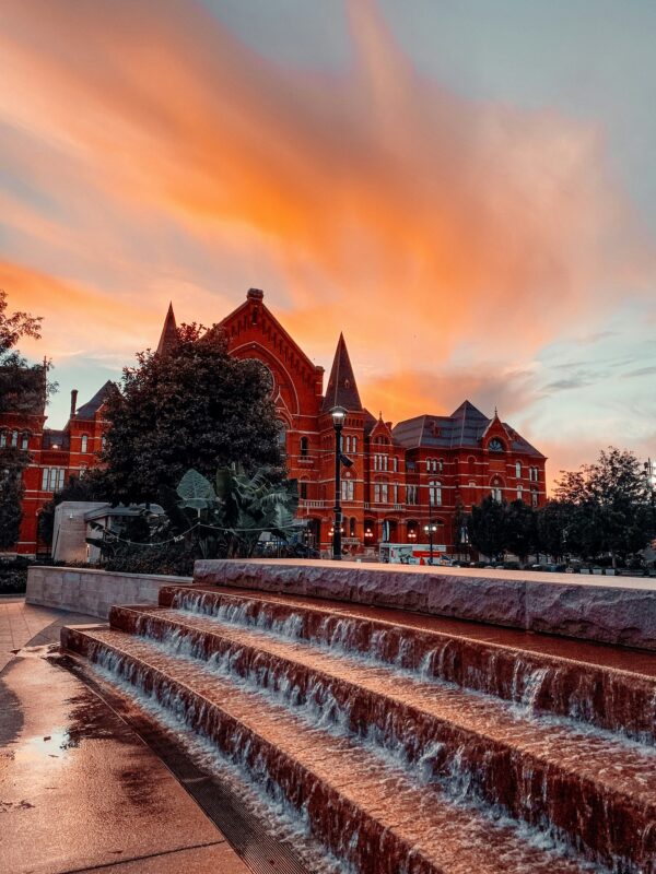 Cincinnati's Music Hall, a Venetian Gothic red brick building from the 1870s. In the foreground, a fountain in Washington Park. Pink clouds drift overhead.