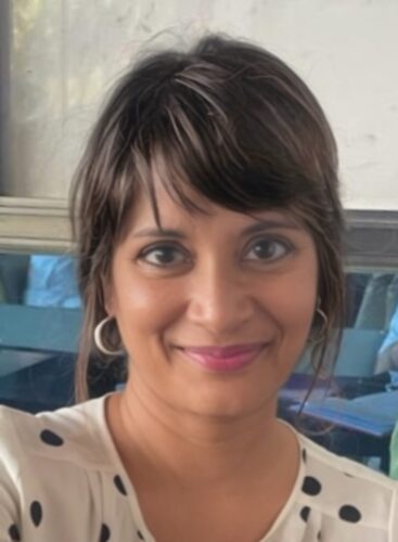 A close headshot of Nandini Bhattacharya, a woman of Desi/Indian background with short black hair. She is wearing hoop earrings and a white shirt with black dots on it. There is a blue booth or couch behind her, and a gray wall.