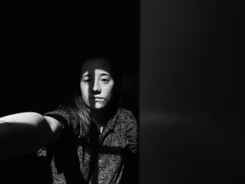 A black-and-white photo of Haley Bossé, a white nonbinary person, in a combination of shadow and light, with their hand out toward the left of the camera. They are looking toward their hand rather than straight at the camera.