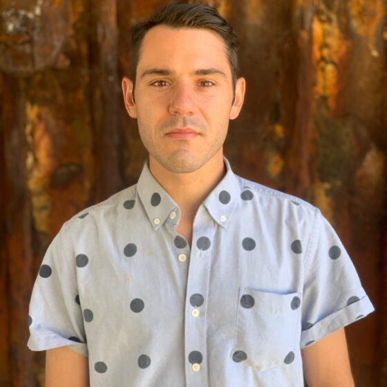 James Ciano, a white man in a short-sleeved blue button down shirt with dark blue polka dots, stands in front of a textured brown background that looks like copper. He has short brown hair and is close-shaved. He doesn't smile.