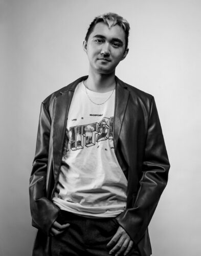 Black-and-white photo of Rob Macaisa Colgate, a multiracial person with short blond and black hair. They are wearing a leather jacket and a graphic T and standing with their thumbs in pant pockets