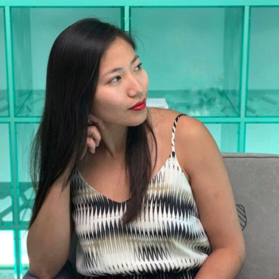 Fran Qi, an Asian-American woman with long hair wearing red lipstick, sits in a chair in front of an illuminated blue background, Her head rests on her hand. She looks to the side. She wears a black-and-white tank top with a sort of wave pattern.