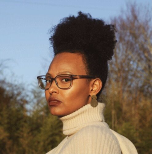 Sadia Hassan, a Somali American woman with a tall ponytail, maroon glasses, and long earrings. She is wearing a white turtleneck and standing outside with blurred sky and trees behind her.