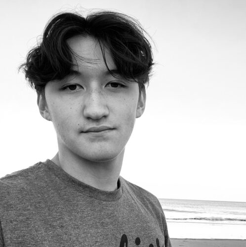 A grayscale picture of Radian Hong in a T-shirt, left of center in the image. There's a large body of water behind him.