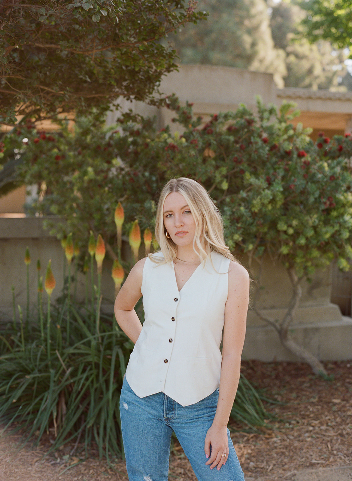 Erika Gallion Velasquez, a white woman with blond hair wearing a tan vest, poses in a garden against a backdrop of trees and flowers.