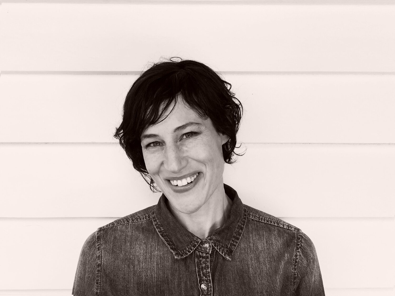 Jaye Kranz, a white woman with short dark hair, wears a denim shirt and smiles in front of a white wooden background.