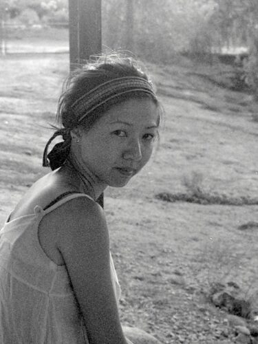 A black-and-white photo of Jennifer S. Cheng, a woman with dark hair held back by an bandana. She is looking at the camera while facing sideways, and she's wearing a light-colored tank top. Behind her is a porch column and a yard.