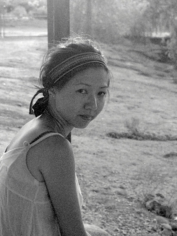 Jennifer S. Cheng, an Asian-American woman, in a black-and-white photo. She wears a white camisole and striped headband with her hair pulled back. She looks over her shoulder at us. She is in front of a grassy natural background.