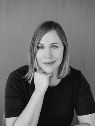 A black-and-white photo of Jessica E. Johnson, a white woman with shoulder-length blond hair. She is wearing a dark short-sleeved shirt and resting her chin on her hand.