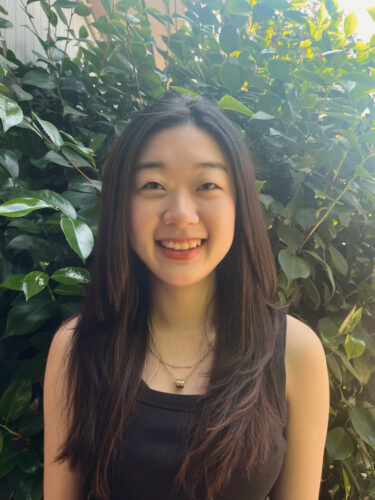 Elane Kim, a Korean American woman with long dark hair, smiling in front of greenery. She is wearing a black tank top and gold necklace with charm.