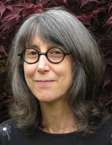 Laurie Blauner, a white woman with shoulder-length gray hair, wears circular black glasses and a black top. She smiles in front of Japanese Maple leaves.