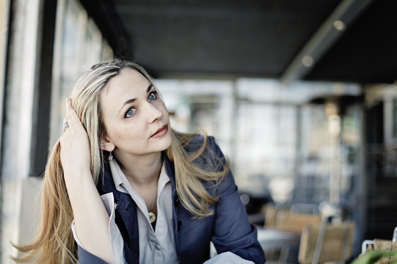 Shara Lessley, a white woman with long blond hair and blue eyes, looking to the right of the image, with her hand in her hair. She's wearing a blue blazer and gray button-up shirt, and there are tables and chairs behind her as if in a cafe.