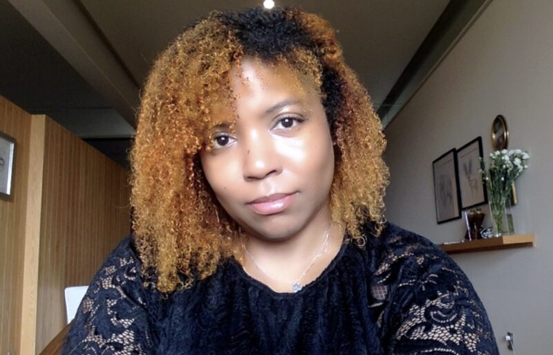 Kimberly Reyes, who identifies as "Black Nuyorican," with reddish and black hair. She is wearing a black shirt with lacy sleeves and a pendant necklace, and there's a narrow room behind her with a shelf of decorations and a light-wood wall.