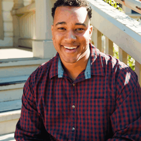 Jalen Eutsey, a young African-American man with short hair and light skin, wears a red-and-black plaid button-down and sits on a staircase with white banisters. He has an open, toothy smile.