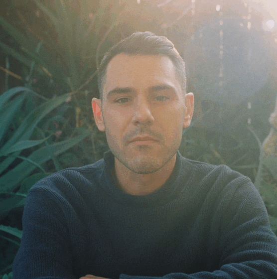 James Ciano, a white man with short, dark hair and a 5-o-clock shadow, wears a dark long-sleeved tee and crosses his arms in front of tropical-looking foliage. The photo has a washed-out, flared quality.