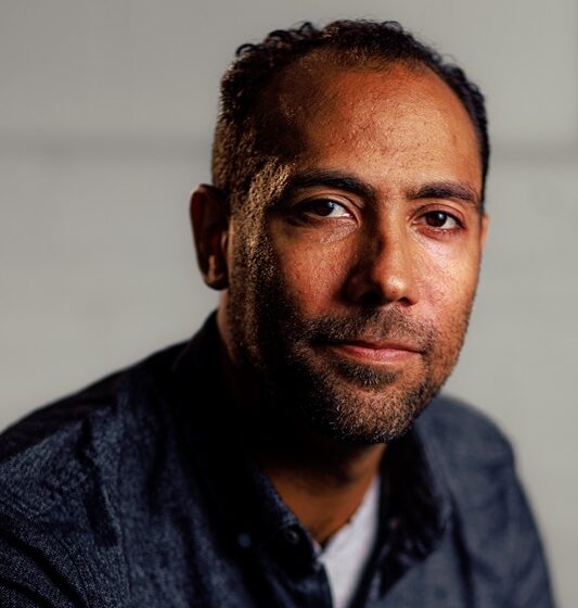 James Stewart III, a Black man with short hair and a five-o-clock shadow, wears a blue chambray shirt and white tee against a gray background. He half-smiles.