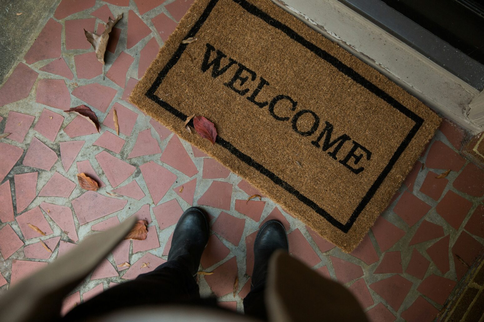 A coir doormat reads "Welcome." Two feet in black shoes stand before it. Dead leaves are scattered around.