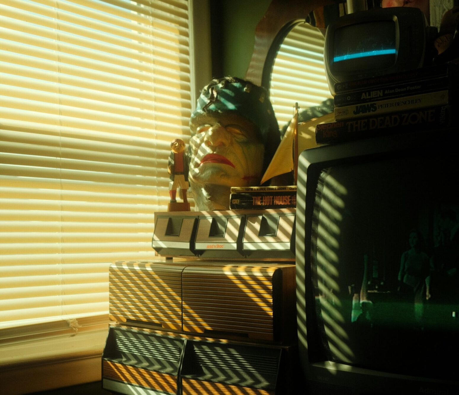 A 90s TV and rubber monster mask sitting on top of some old cabinets next to a window with its blinds shut against the afternoon sunlight.