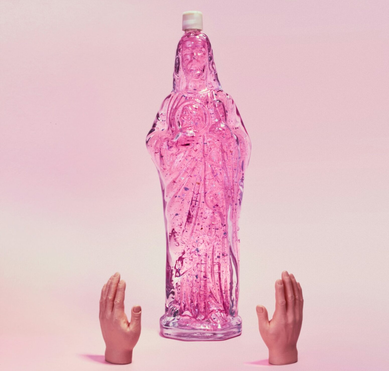 A bottle of sparkly bubble bath in the shape of the Virgin Mary with two small plastic hands set in front of it as if they're raised in prayer.