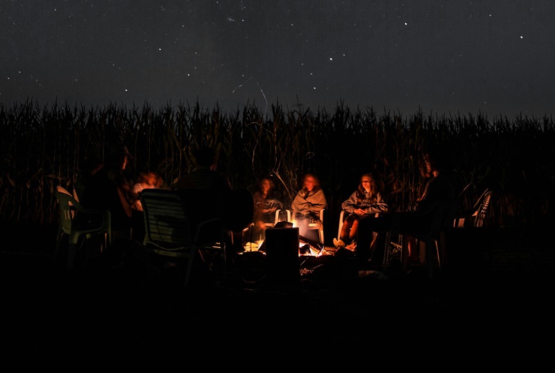 A group of people gathered around a campfire at night with a field behind them and stars above.