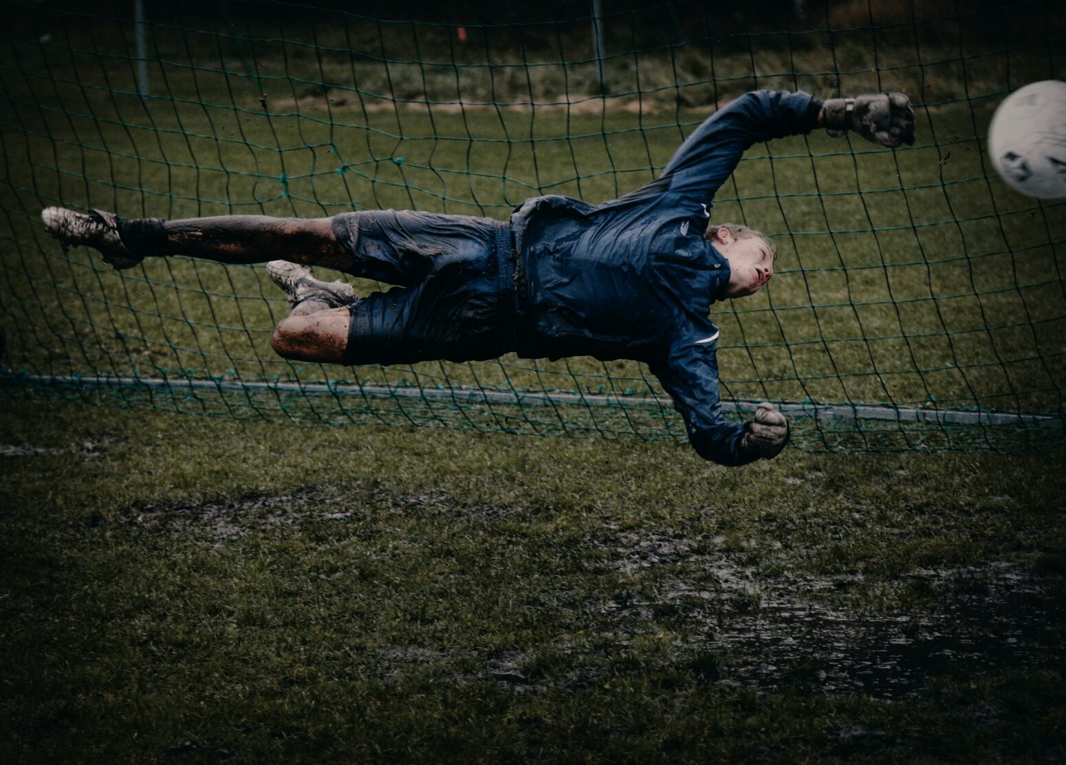 A soccer goalkeeper is caught midair, perfectly parallel to the muddy ground. He has an arm outstretched to block the ball, and a leg outstretched like a dancer. One of his legs kicks behind him.