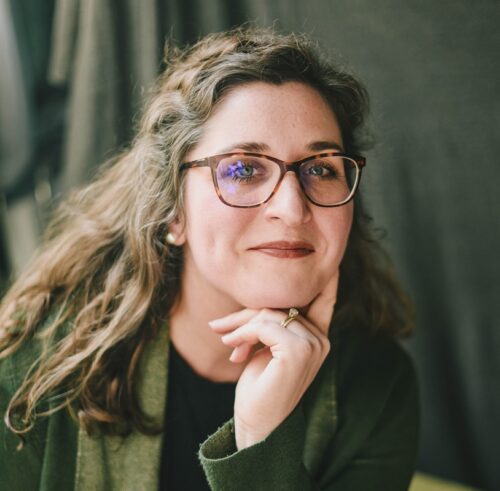 Mary Ardery, a white woman with curly brown hair past her shoulders. She is wearing tortoiseshell glasses and lipstick, and she has her left hand tucked near her chin. She's wearing a green scarf and jacket.