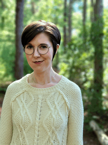 Sarah Fawn Montgomery, a white woman with short brown hair in a pixie cut. She is wearing a white cable-neck sweater and glasses, and smiling as she stands amid trees.