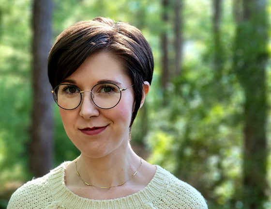 A woman with a dark, brown pixie cut, smiling. She stands in front of a forest background, wearing a yellow sweater and round, wire glasses.