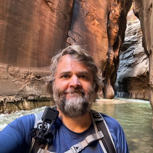 Jason Gray, a white man with gray and black hair and beard. He is wearing a dark blue shirt and backpack and standing in a rock formation with water.