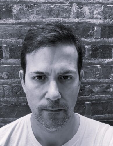 A black-and-white headshot of Shane Joaquin Jimenez, a Latino man with dark hair and a light goatee, standing in front of a bright wall.