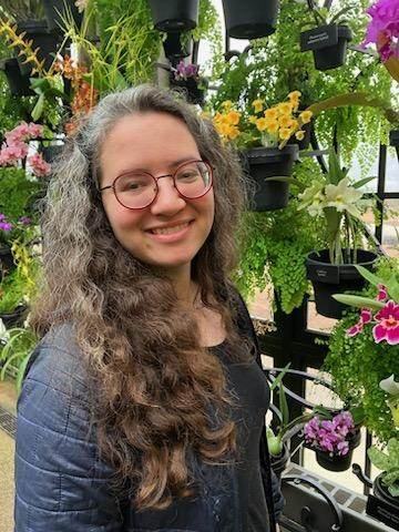Louise Ling Edwards, a biracial woman with brown and gray curly hair. She is wearing red glasses, smiling, and standing in front of an array of potted plants.
