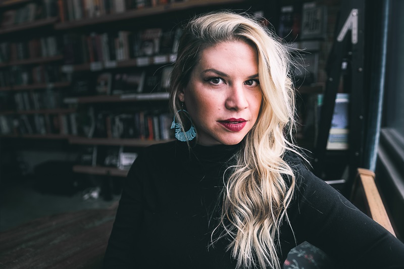 Jenny Molberg, a white woman with long blond hair arranged in waves over her left shoulder. She is wearing large blue hoop-style earrings and a black top and is seated in a darkened room with bookshelves by a window.