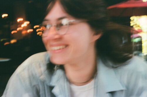 A blurry photo of Gabrielle Grace Hogan, a white woman with dark hair and glasses, smiling. She's wearing a collared shirt over a T-shirt, and there are blurred lights behind her.