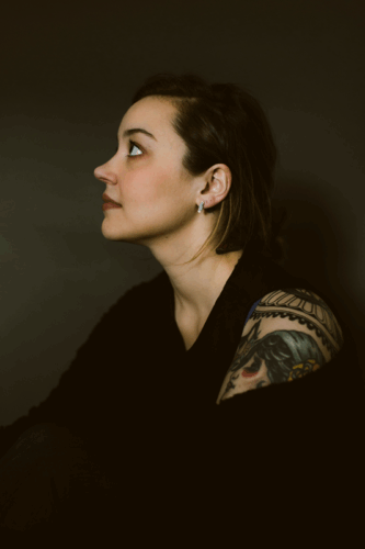 Brenna Womer, a Latine person with she/they pronouns, in profile. She has short dark hair and a colorful tattoo on her left shoulder. She's wearing all black against a dark-gray blackground and looking to the left.