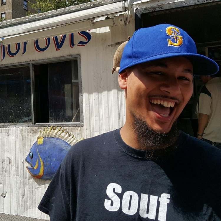 Malcolm Friend, a Black man with a beard and mustache wearing a blue Mariners cap and black tee that reads "Souf," stands in front of a tin shed with a blue metal fish on its side.
