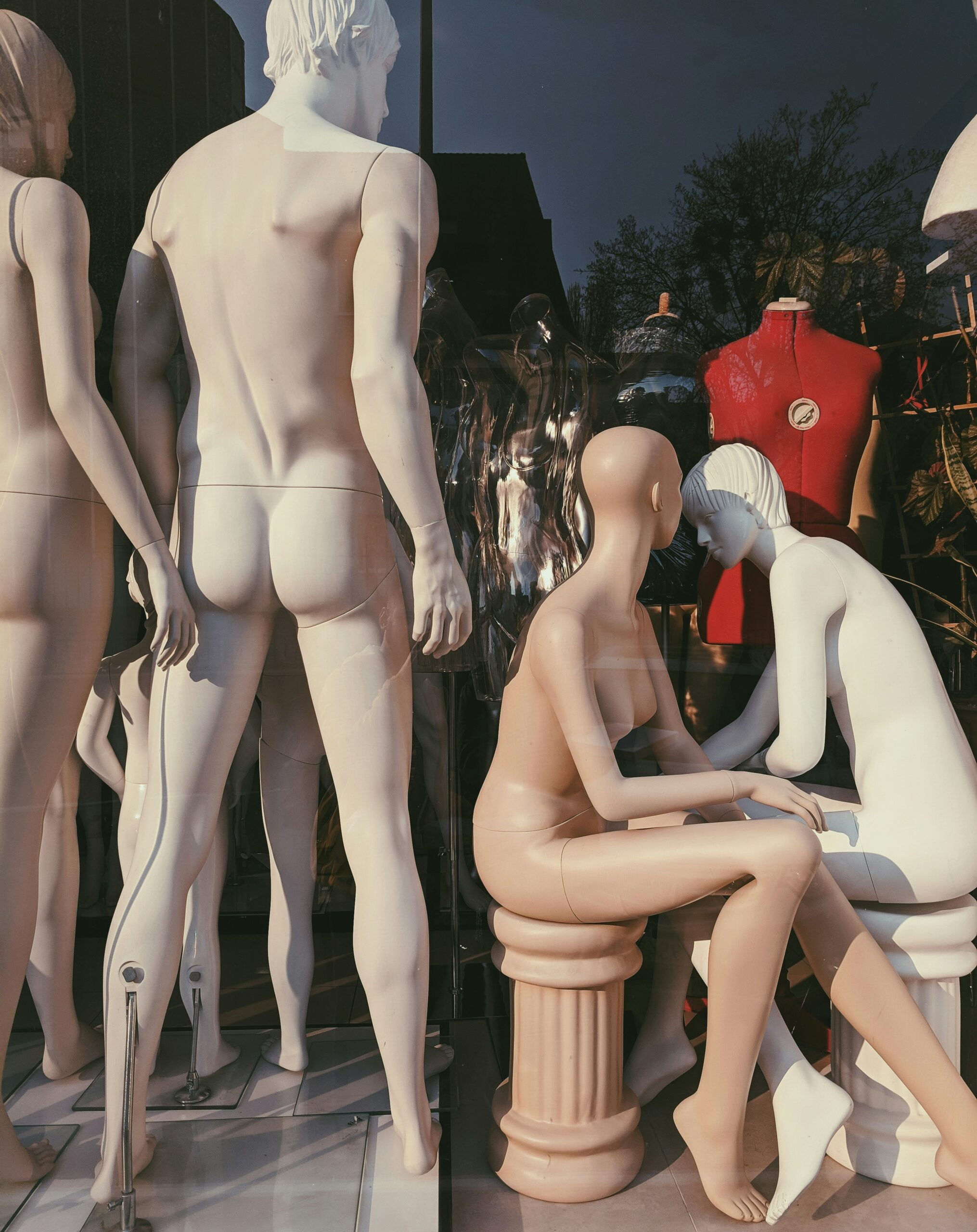 Male and female mannequins in a window display. They are all facing away from us; the male mannequin has a very shapely butt. Two of the female mannequins seem to be sitting on low columns and conversing with each other.